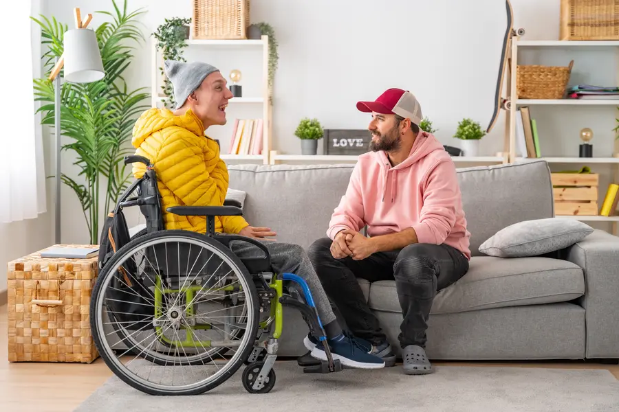 Young man in wheelchair inn his living room enjoying a conversation with his male support worker