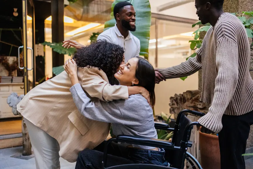Girl in wheelchair hugging a female support worker while two male friends shake hands in the background